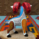 This Oaxacan ram wood carving by Abundia Munoz features large red curled horns, purple ears, a white body, and intricate black, orange, brown, and turquoise geometric patterns; side/back facing view.