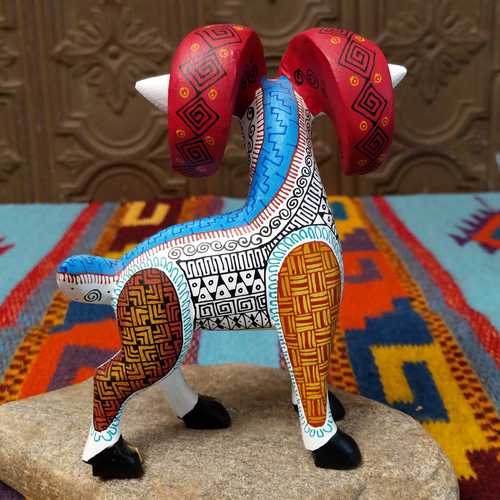 This Oaxacan ram wood carving by Abundia Munoz features large red curled horns, purple ears, a white body, and intricate black, orange, brown, and turquoise geometric patterns; side/back facing view.
