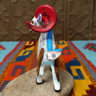 This Oaxacan ram wood carving by Abundia Munoz features large red curled horns, purple ears, a white body, and intricate black, orange, brown, and turquoise geometric patterns front/side facing view.