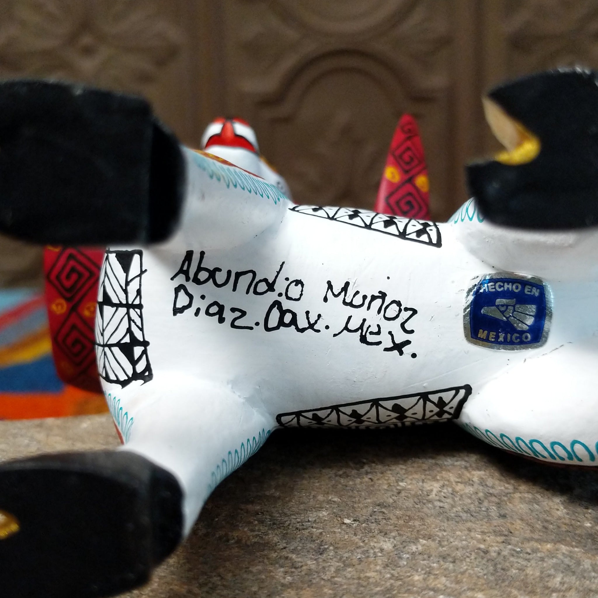 This Oaxacan ram wood carving by Abundia Munoz features large red curled horns, purple ears, a white body, and intricate black, orange, brown, and turquoise geometric patterns; artisan and production location.