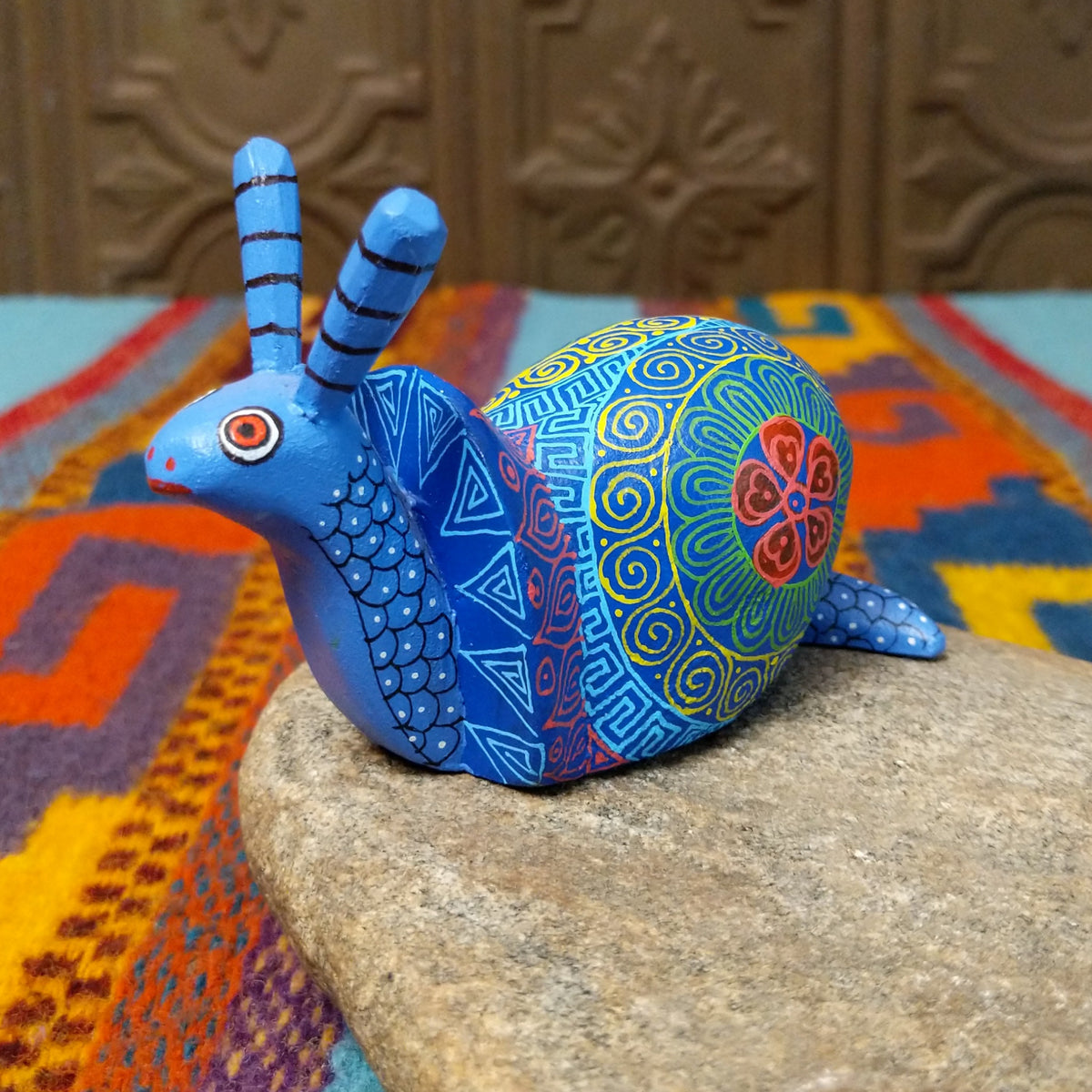 This blue Oaxacan snail wood carving by Alberto Hernandez Melchor features tall striped antennae and a colorful shell decorated with intricate spirals, geometric patterns, and a floral motif.
