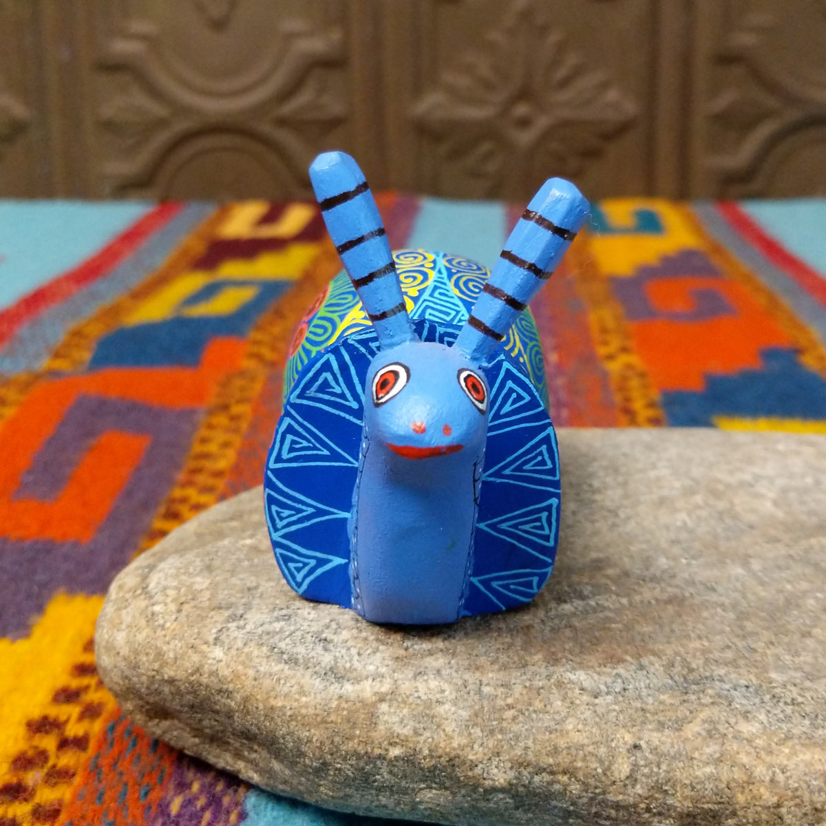 This blue Oaxacan snail wood carving by Alberto Hernandez Melchor features tall striped antennae and a colorful shell decorated with intricate spirals, geometric patterns, and a floral motif; front view.