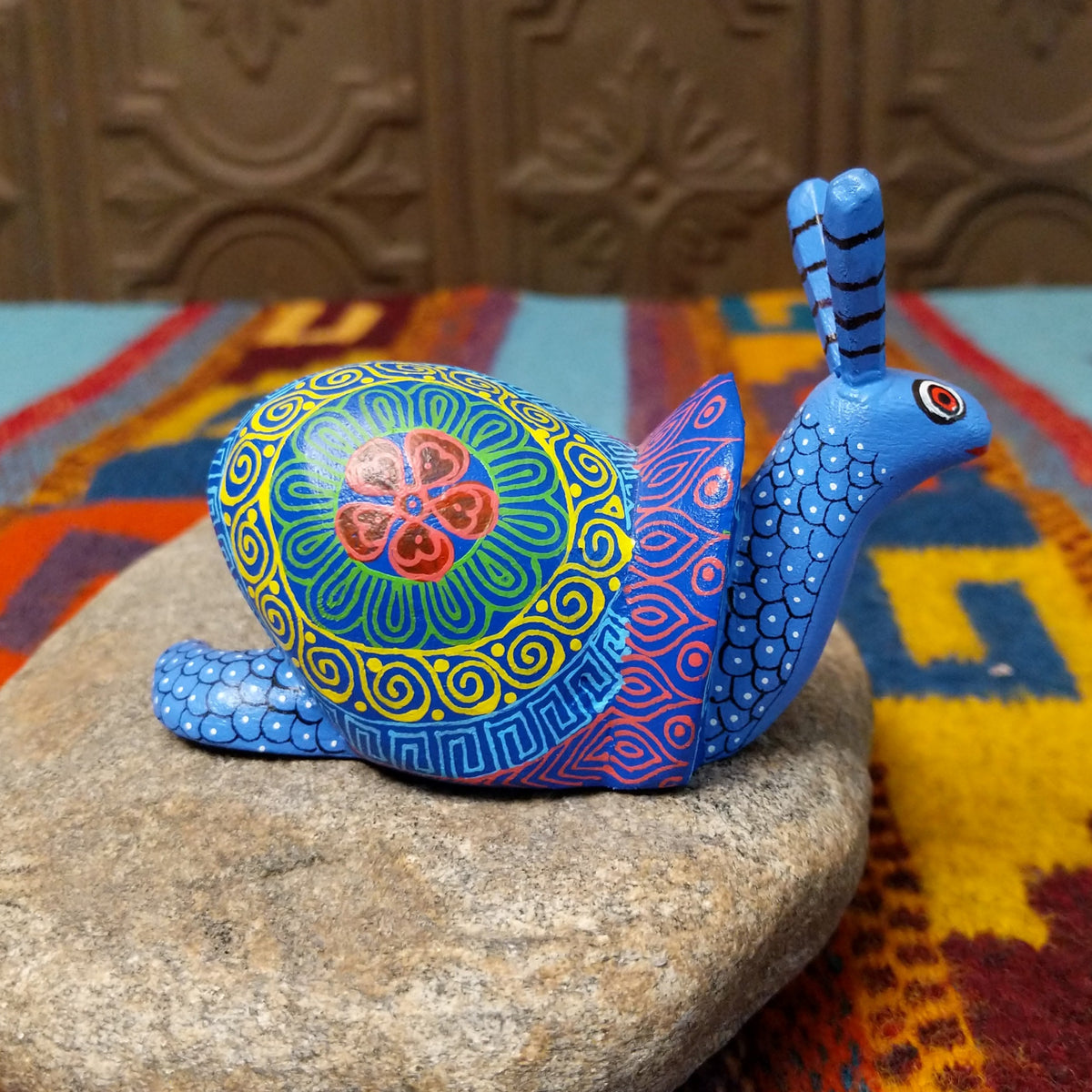 This blue Oaxacan snail wood carving by Alberto Hernandez Melchor features tall striped antennae and a colorful shell decorated with intricate spirals, geometric patterns, and a floral motif; side view.