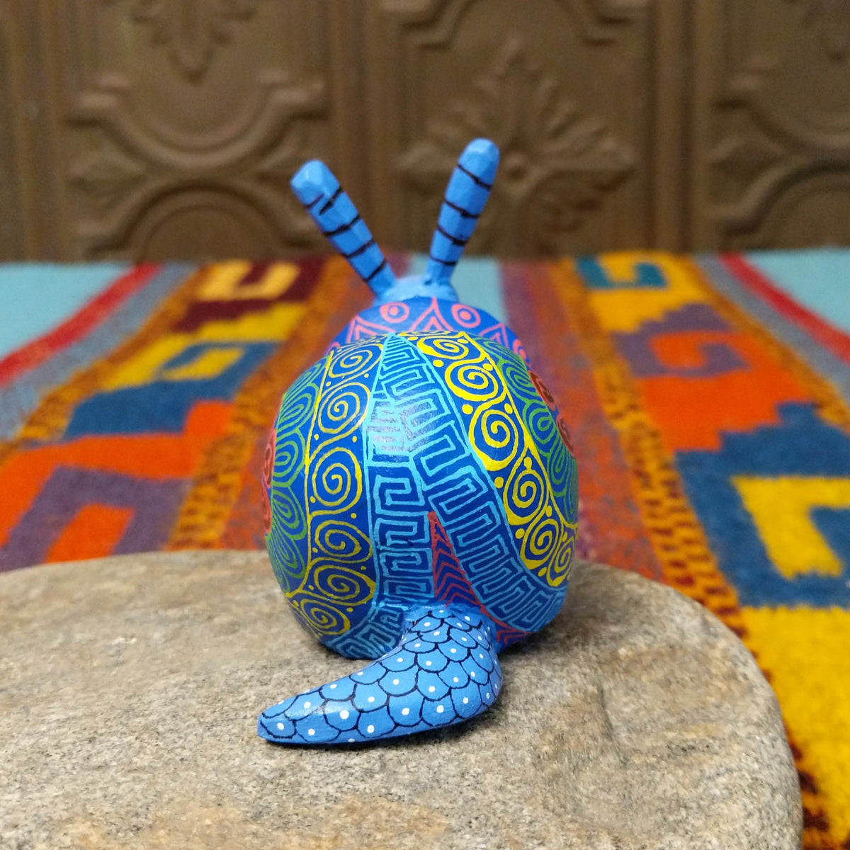 This blue Oaxacan snail wood carving by Alberto Hernandez Melchor features tall striped antennae and a colorful shell decorated with intricate spirals, geometric patterns, and a floral motif; rear view.