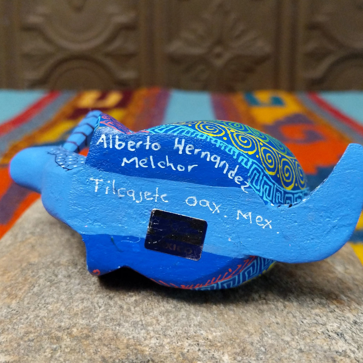 This blue Oaxacan snail wood carving by Alberto Hernandez Melchor features tall striped antennae and a colorful shell decorated with intricate spirals, geometric patterns, and a floral motif; artisan and made in Mexico signs.