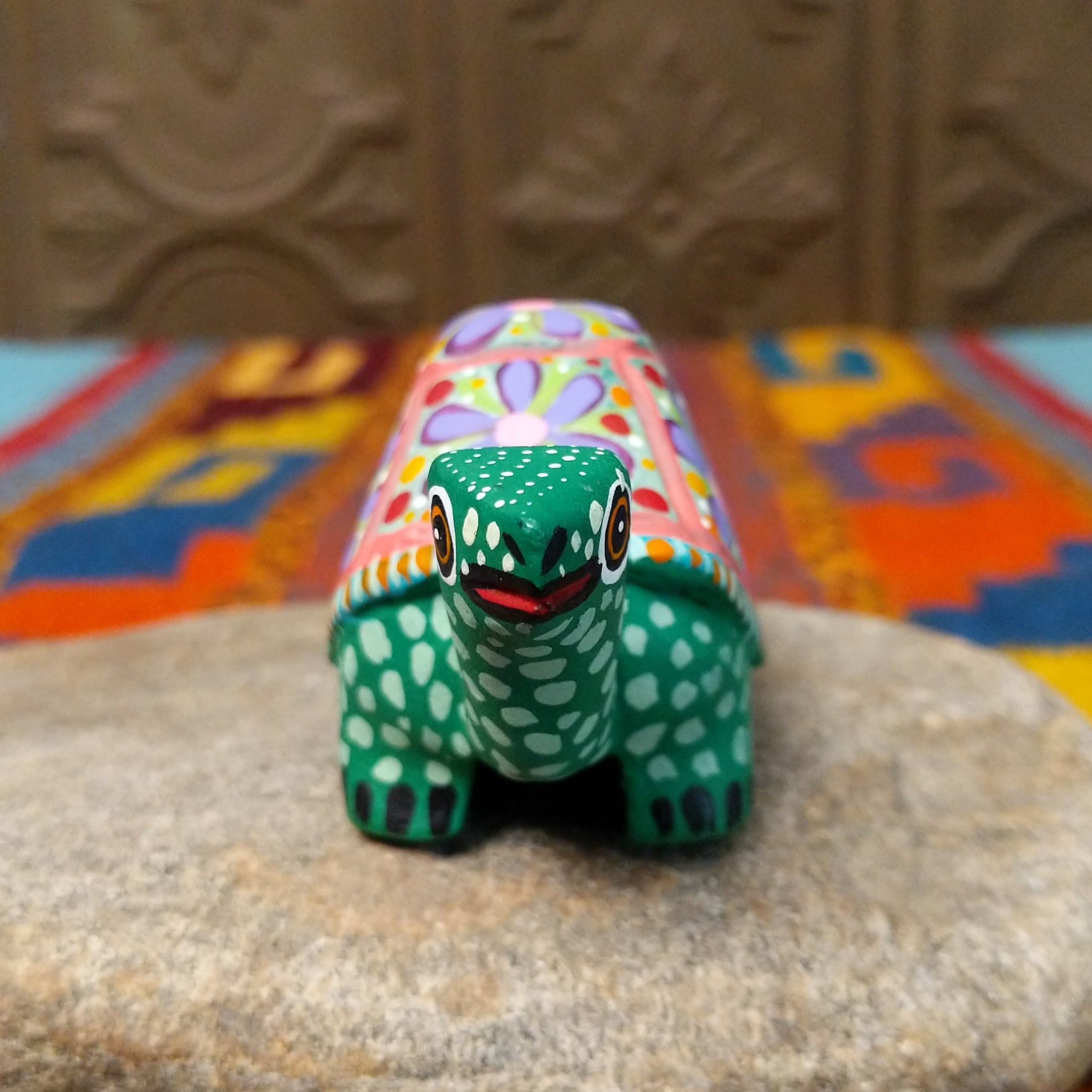 This green Oaxacan tortoise wood carving by Rosaura Ortega features a domed shell decorated with purple flowers, pink borders, and colorful hand-painted dots and patterns; front facing view.