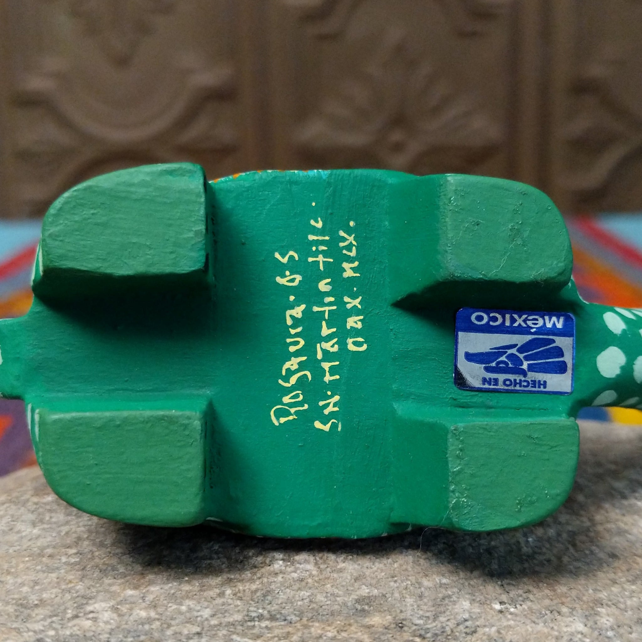 This green Oaxacan tortoise wood carving by Rosaura Ortega features a domed shell decorated with purple flowers, pink borders, and colorful hand-painted dots and patterns; artisan and production location written on underside.