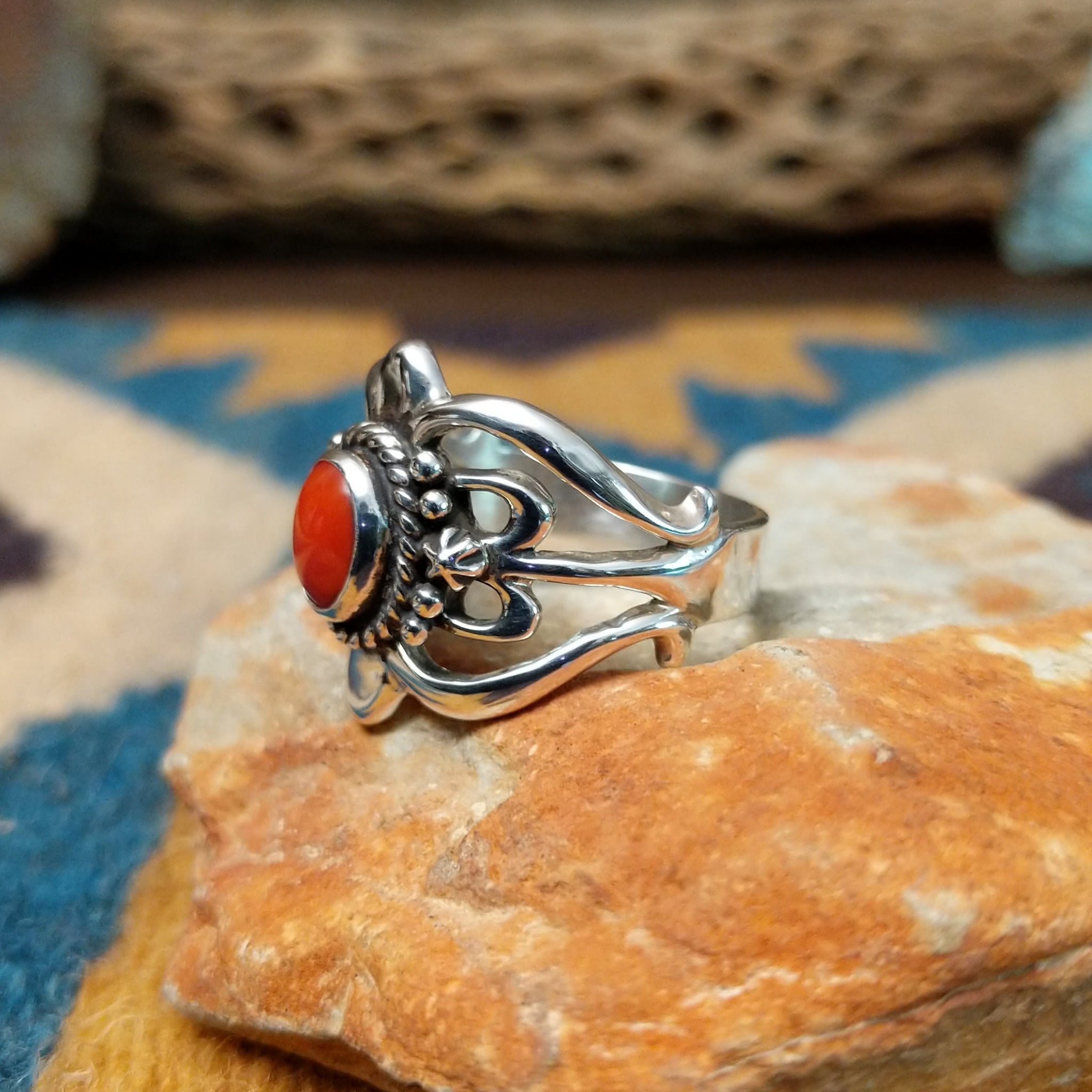 Handcrafted Navajo sterling silver ring by Raymond Coriz featuring an oval red coral cabochon surrounded by twisted rope, silver bead accents, and an ornate openwork floral design; side view.