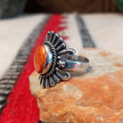 Ornate red spiny oyster shell ring by Raymond Coriz featuring an oval red-orange shell cabochon surrounded by twisted rope, fan-shaped silverwork, scrolls, and decorative silver beads; side view.
