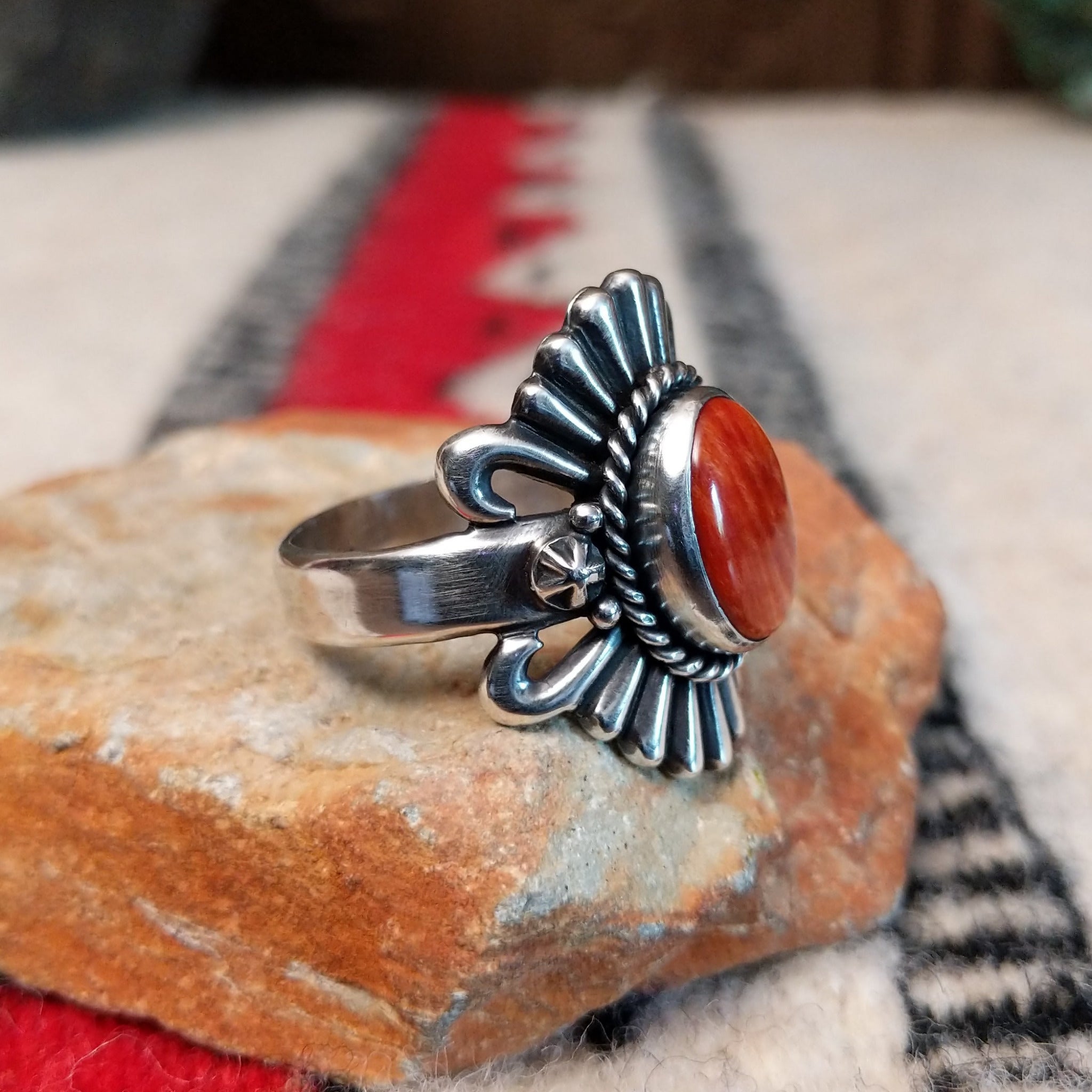 Ornate red spiny oyster shell ring by Raymond Coriz featuring an oval red-orange shell cabochon surrounded by twisted rope, fan-shaped silverwork, scrolls, and decorative silver beads; side view.