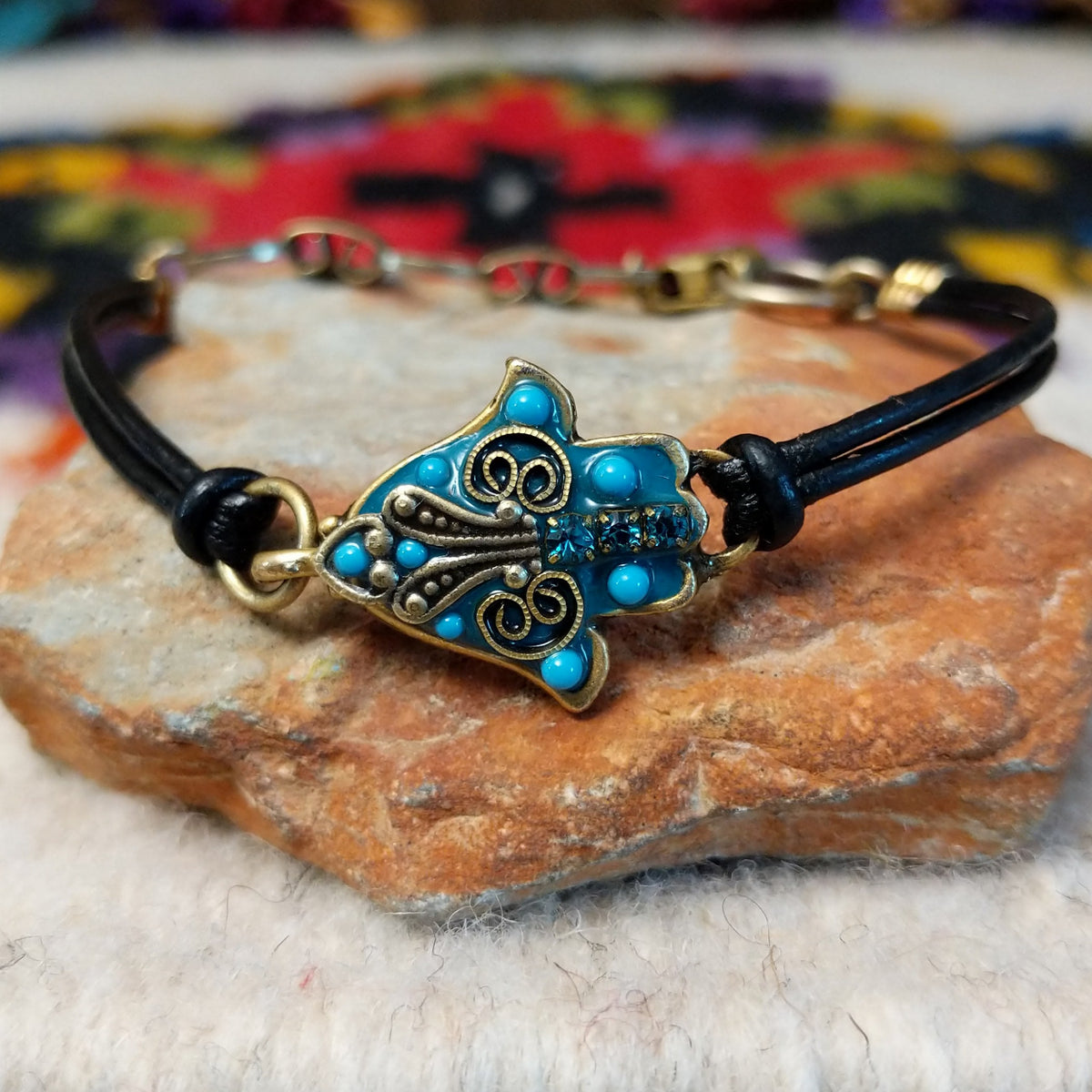 Ornate Hamsa bracelet with a turquoise-colored centerpiece, antique-gold scrollwork, blue crystal accents, and a double-strand black leather cord.