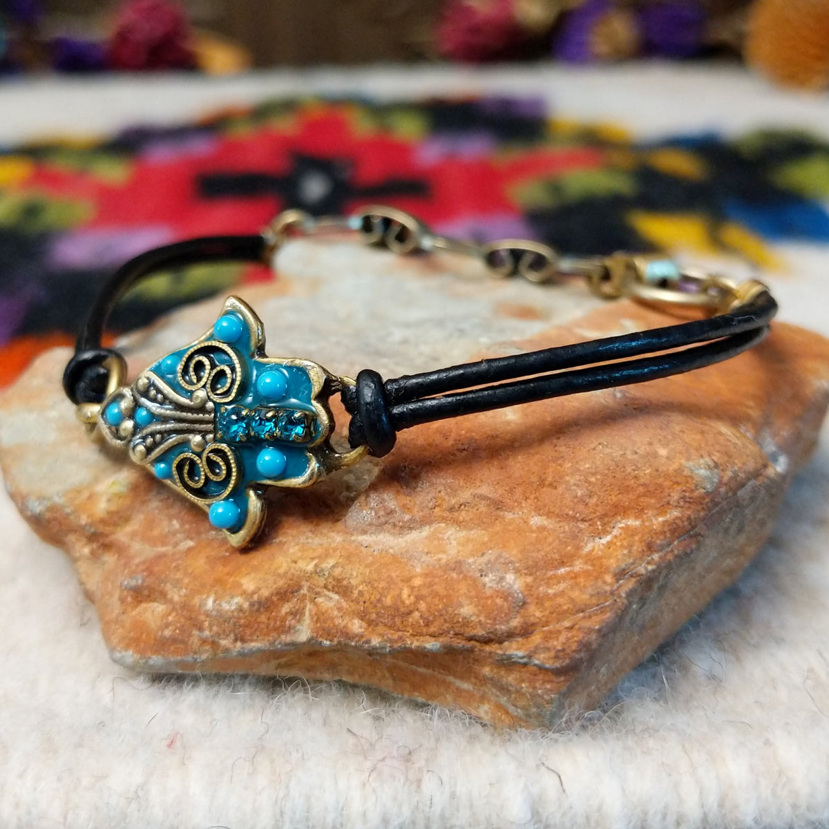 Ornate Hamsa bracelet with a turquoise-colored centerpiece, antique-gold scrollwork, blue crystal accents, and a double-strand black leather cord; side view.