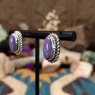 Handcrafted sterling silver post earrings by Priscilla Smith featuring oval charoite cabochons in polished bezel settings with traditional twisted rope border detailing; side view.