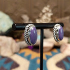 Handcrafted sterling silver post earrings by Priscilla Smith featuring oval charoite cabochons in polished bezel settings with traditional twisted rope border detailing; side view.