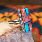  Size 10 ring by Wilbert Grey showcases an inlay design set in sterling silver. The rectangular face features a striking mosaic of vibrant stones, including rich red and orange spiney oyster shell, bright turquoise, and deep blue lapis. Side view