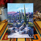 Party Cactus canvas artwork by Diana Madaras featuring tall green saguaro cacti, prickly pear plants, desert rocks, yellow wildflowers, and purple mountains beneath a blue Arizona sky.