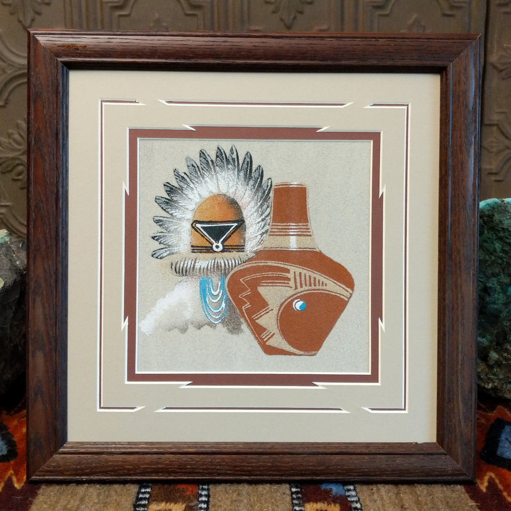 Multiple colored sand used to depict a kachina and pot set within a wooden frame.