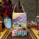 Prickly Pear Club Bag by Bamboo Trading featuring a colorful beaded Southwestern desert landscape with purple mountains, pink and yellow sunset sky, prickly pear cacti with coral-colored blossoms, desert plants, sparkling sequins, and a cream-colored shoulder strap.
