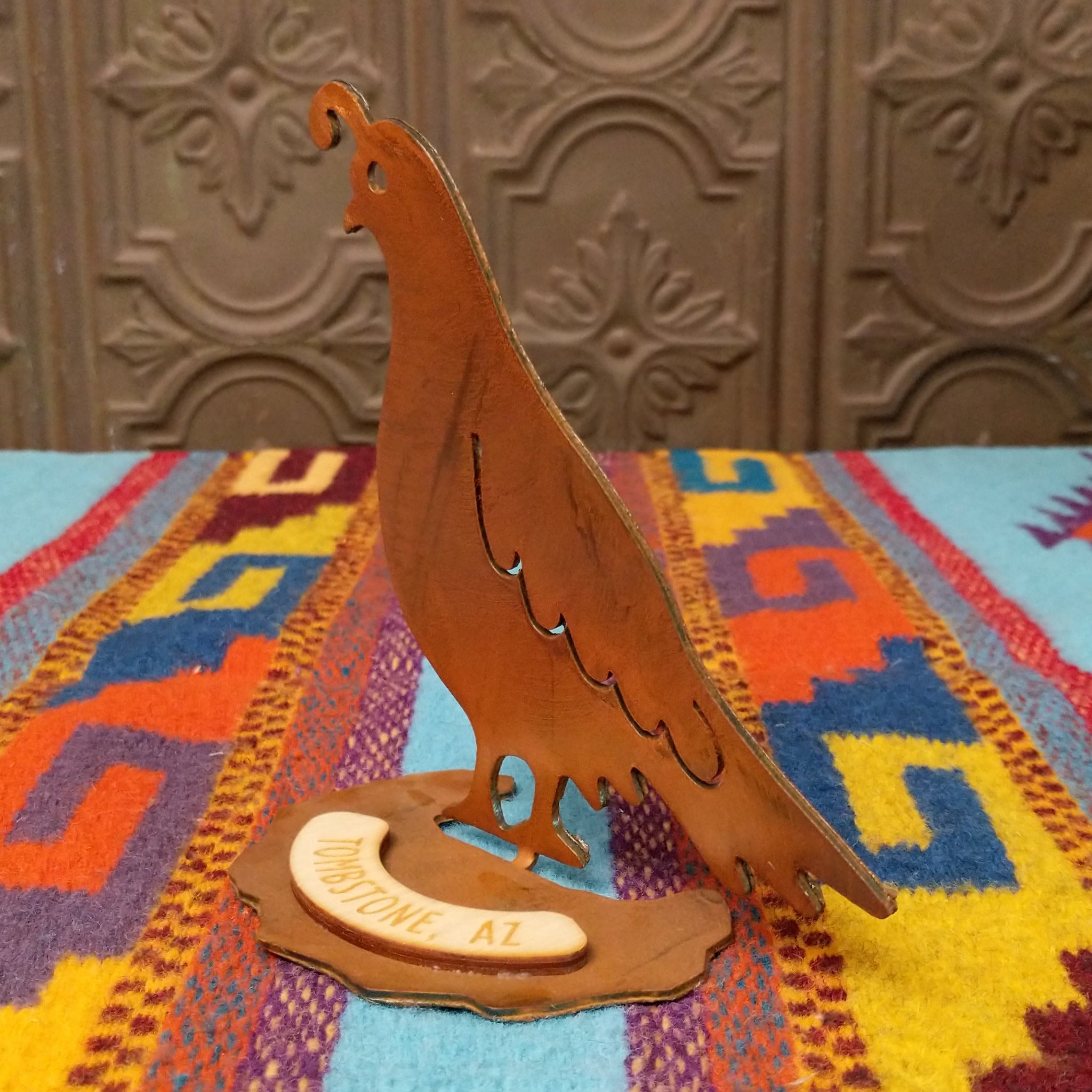 Handcrafted Crooks Design metal quail tabletop sculpture with a curved topknot, cutout wing details, warm rustic finish, and decorative base marked Tombstone, AZ; side view.