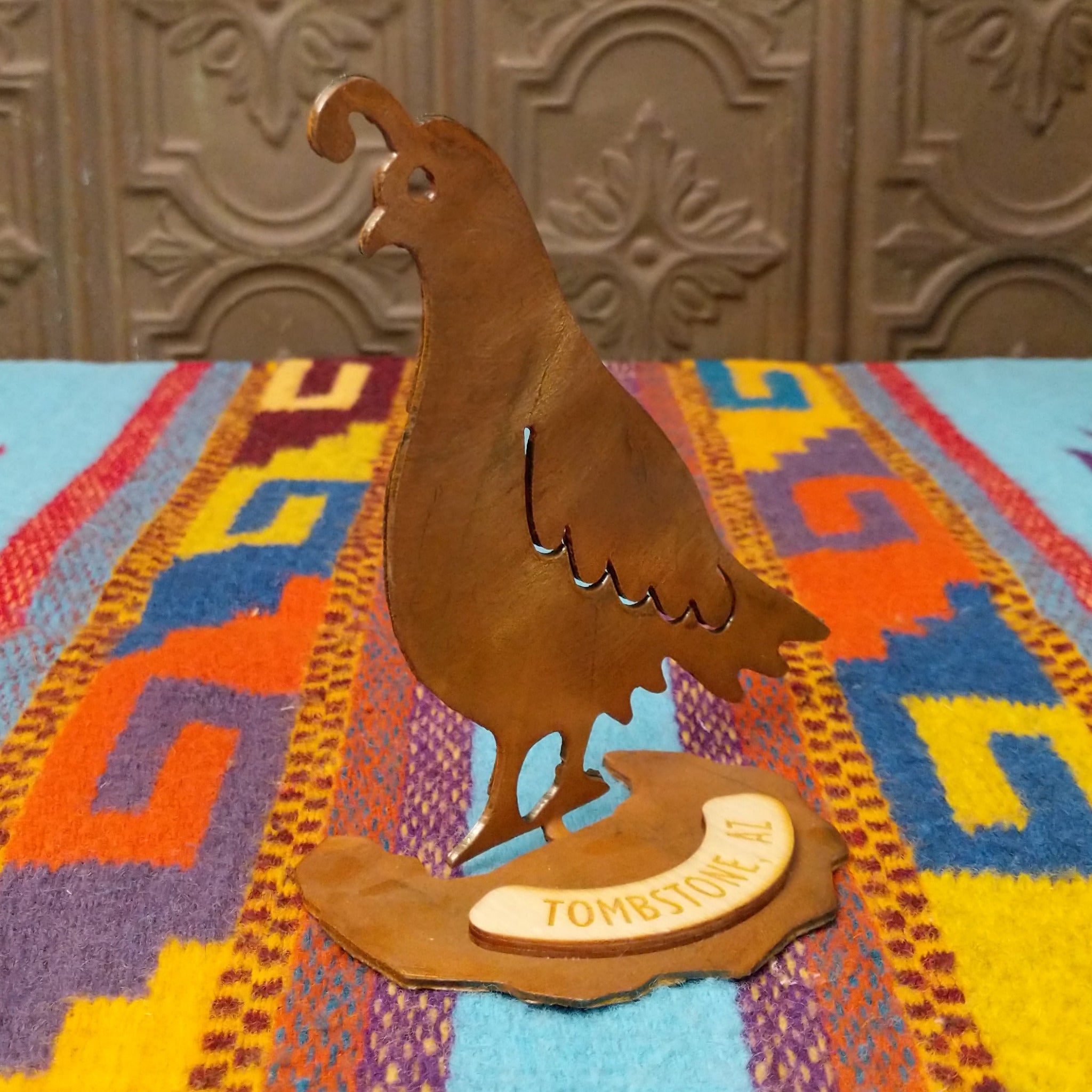 Handcrafted Crooks Design metal quail tabletop sculpture with a curved topknot, cutout wing details, warm rustic finish, and decorative base marked Tombstone, AZ; side view.