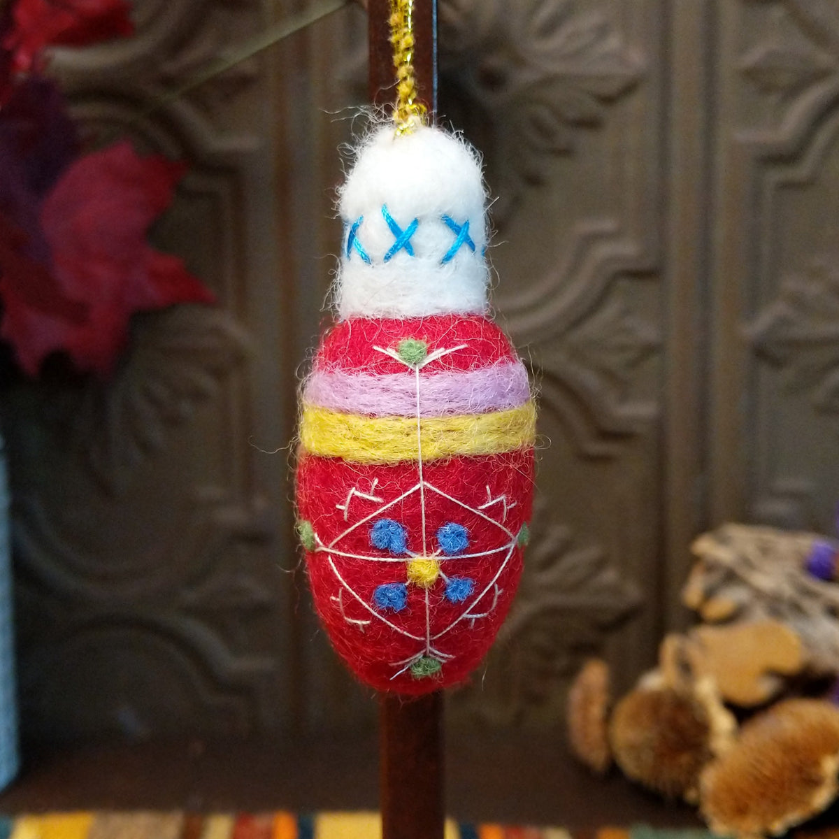 This charming Red Bulb Wool Ornament by Ganz features a bright red wool body with white embroidered geometric designs, blue and yellow floral accents, lavender and yellow bands, a fluffy white top with blue stitching, and a gold hanging cord.
