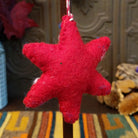 This charming Red Star Wool Ornament features a vibrant red six-pointed star with embroidered green branches, a white flower with a yellow center, soft pink floral accents, blue details, and a red and white twisted hanging cord; back side view.