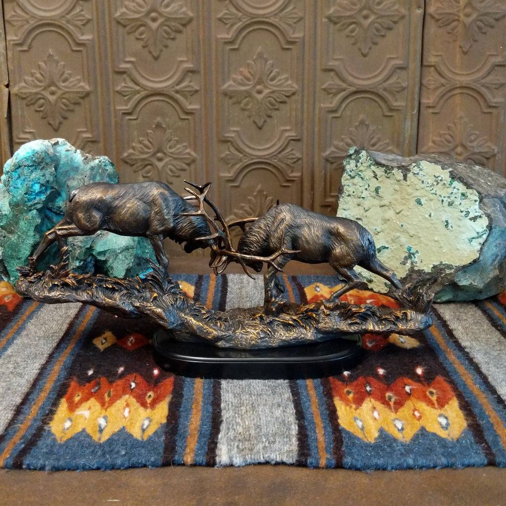 Sculpture showing two bronze stags with antlers locked in combat on a textured base.