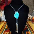 A turquoise stone bolo tie necklace with a black braided cord and Sterling Silver dangling tips, displayed on a black velvet mannequin bust.
