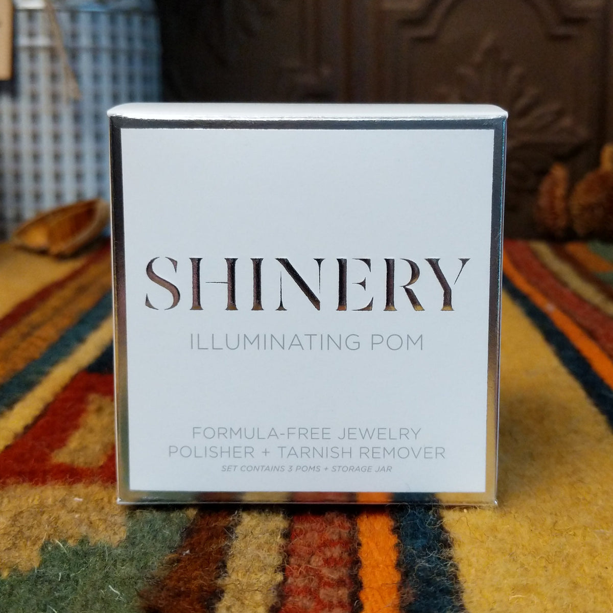 A square white box of Shinery Illuminating Pom jewelry polish and remover, standing upright on a patterned yellow, orange, and green rug with a decorative brown background.
