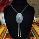 A silver-toned oval bolo tie with an engraved floral center, suspended on a braided black cord, displayed on a black velvet mannequin bust.