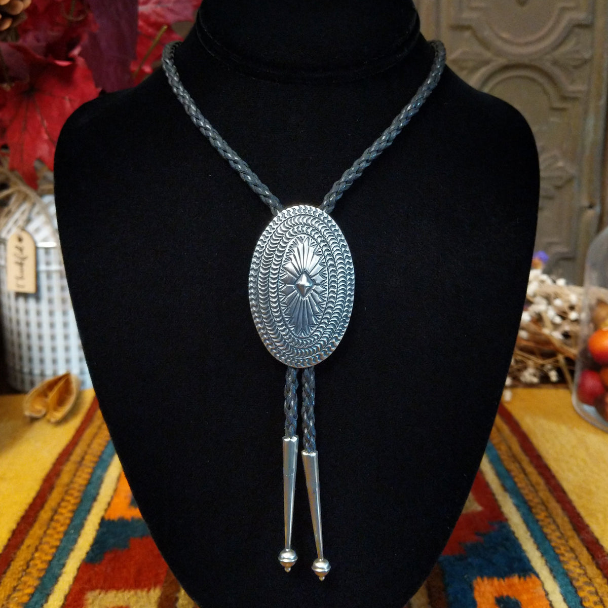 A silver-toned oval bolo tie with an engraved floral center, suspended on a braided black cord, displayed on a black velvet mannequin bust.