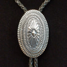 A silver-toned oval bolo tie pendant with intricate engraved patterns and a braided cord, displayed against a black background.