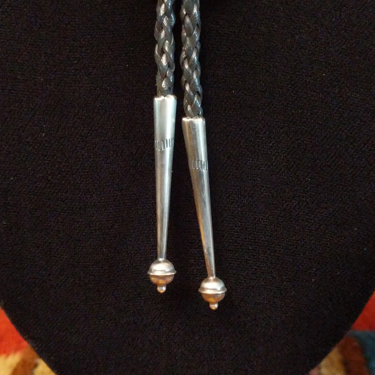 A close-up of a silver bolo tie with braided black cords, displayed on a black velvet jewelry bust.
