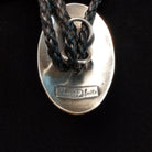 A close-up of  the back of the silver oval bolo tie with a black braided leather cord, featuring a brand engraving that reads 'Chanel De Kuita' on a black background.