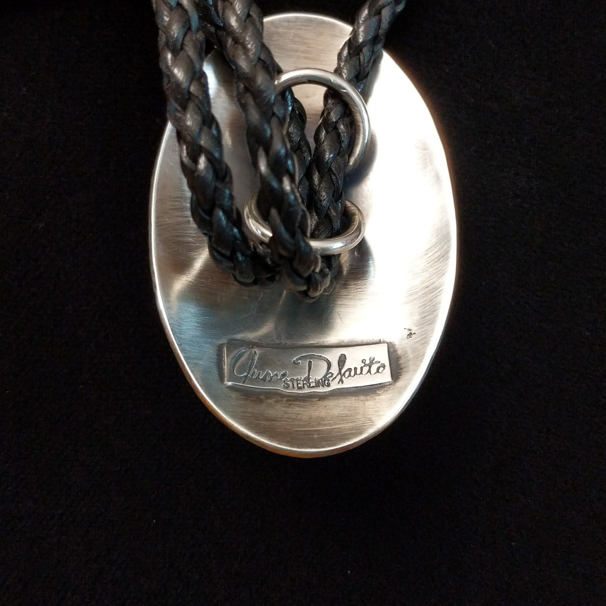 A close-up of  the back of the silver oval bolo tie with a black braided leather cord, featuring a brand engraving that reads 'Chanel De Kuita' on a black background.