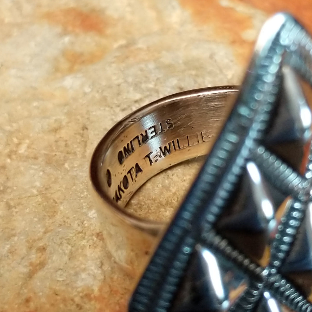Silver ring by Dakota T. Willie featuring an elongated diamond-shaped face with nine raised, polished studs arranged within textured geometric borders. Close up of the artist's stamped signature.
