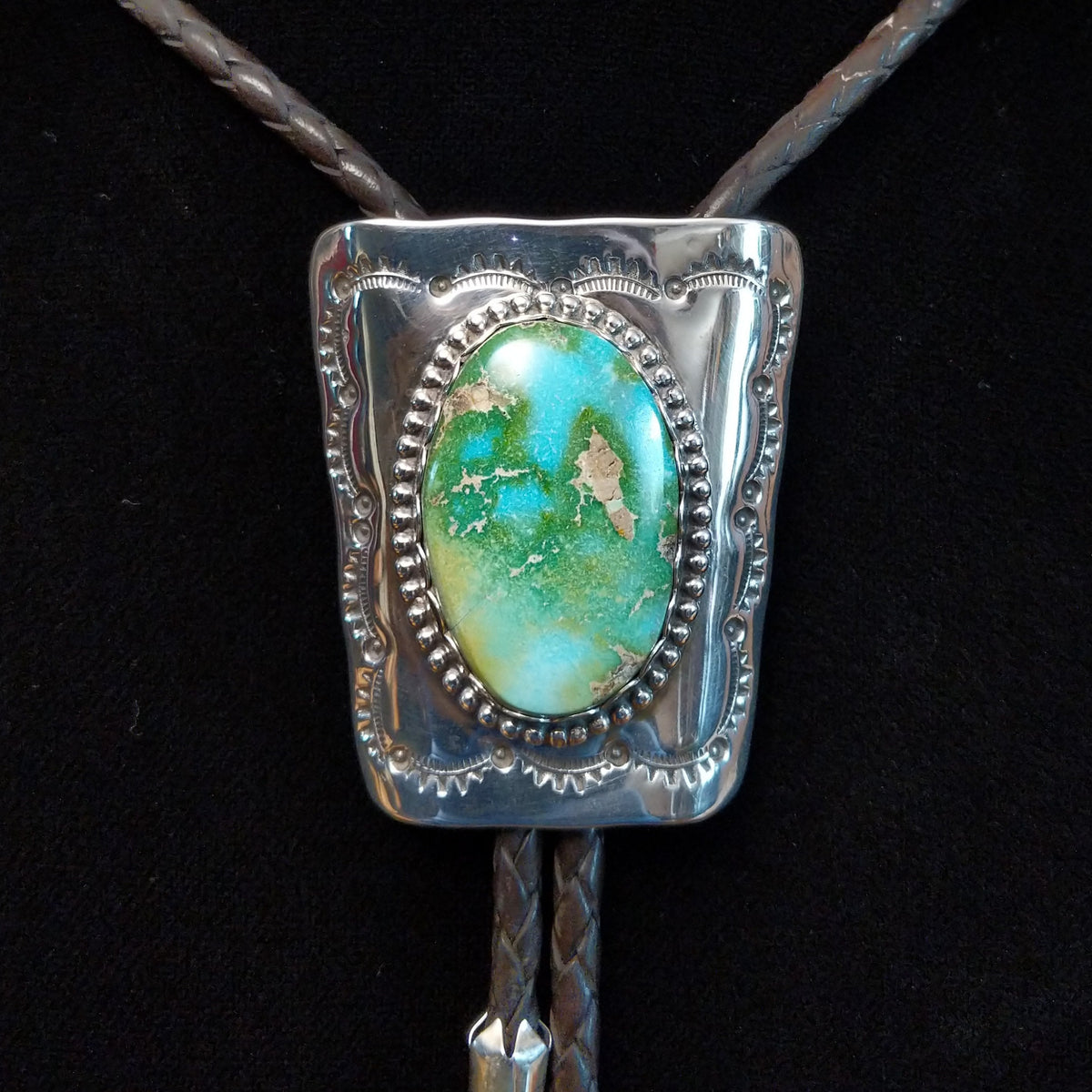 A sterling silver bolo tie necklace featuring a rectangular silver pendant with a Sonoran Gold Turquoise stone, suspended from braided black rope on a black background.
