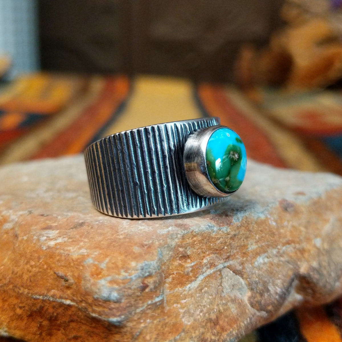 Sonoran Gold Turquoise Ring by Elgin Tom featuring a round blue and green turquoise stone with natural matrix, set in a wide sterling silver band with textured vertical lines; side view.