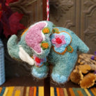 This charming Spearmint Colored Elephant Wool Ornament features a soft green body, pink ears, golden yellow hair, white tusks, blue feet, colorful embroidered details, and a red and white twisted hanging cord.