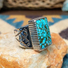 Spiderweb Kingman Turquoise Ring by Joe Piaso Jr. featuring a large rectangular sky-blue Kingman turquoise stone with an intricate dark spiderweb matrix, set in a sterling silver serrated bezel with an openwork silver band, displayed on a natural stone; side view.