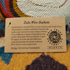 Handwoven square wire art bowl by Bridge for Africa featuring a spiral pattern in recycled wire with ivory, rust, blue, gold, brown, and charcoal tones, ideal for home décor or wall display; artisan information.