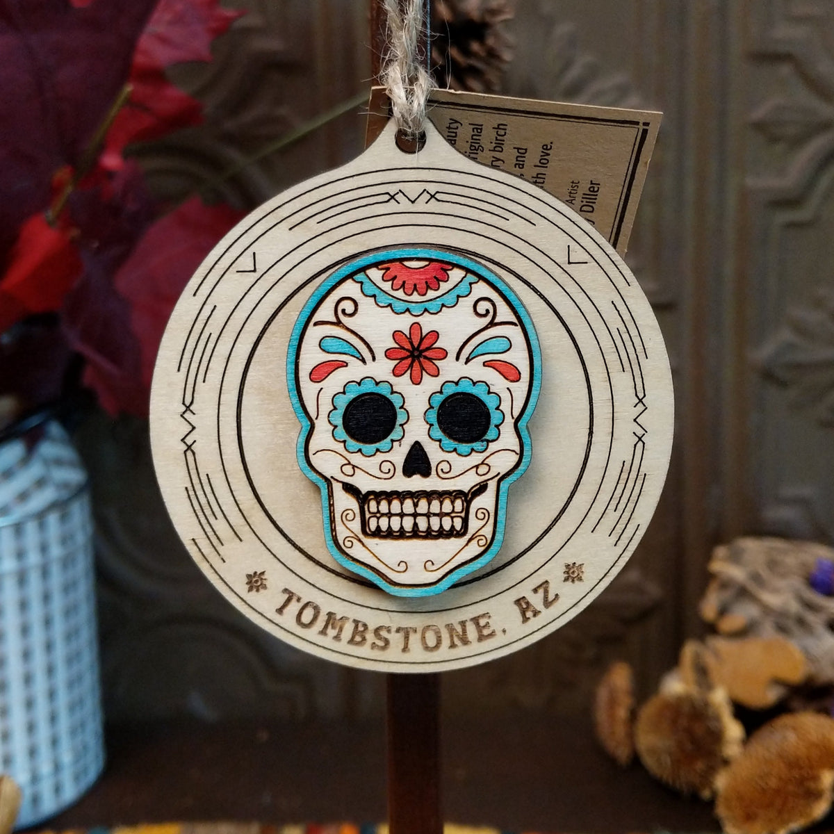 Sugar Skull Wood Ornament featuring a colorful floral skull with turquoise blue and red accents, engraved circular wooden frame, “Tombstone, AZ” lettering, and natural twine hanging loop.