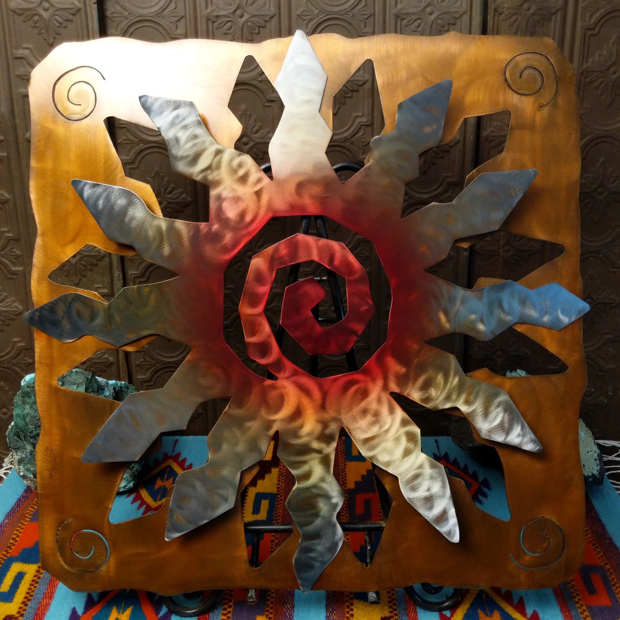 This Crooks Design metal wall art features a 12-point sunburst with angular silver and gold rays, a deep red spiral center, and a warm golden-brown frame with spiral accents.