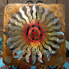 This Crooks Design metal wall art features a 24-point sunburst with flowing silver and gold rays, a red and orange spiral center, and a warm golden-brown frame with spiral accents.