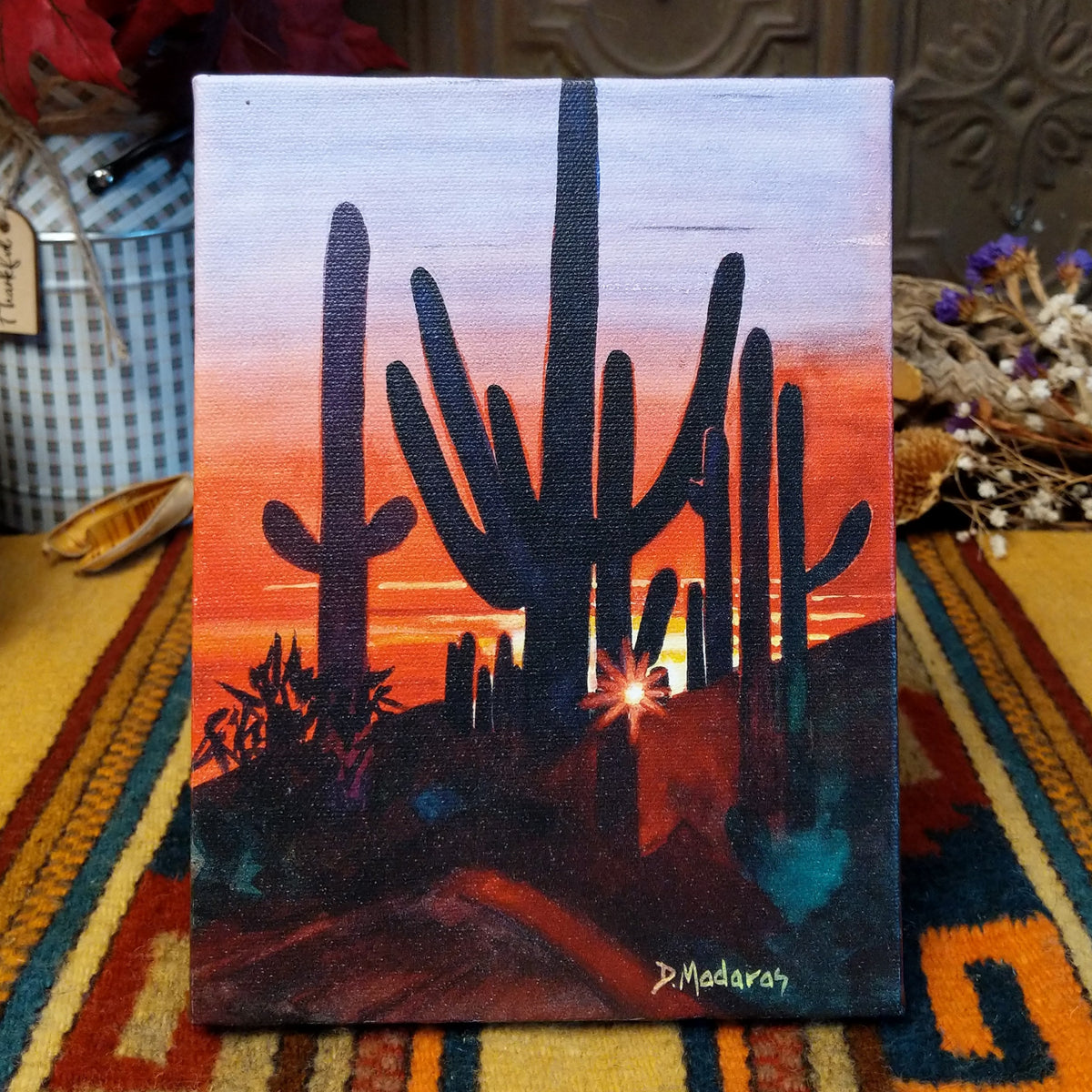 Sunset at Dove Mountain canvas artwork by Diana Madaras featuring dark saguaro cactus silhouettes against a vibrant orange, red, and purple desert sunset with the sun glowing near the horizon.