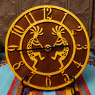 This Crooks Design round Tableau Clock features a pair of golden-toned Kokopelli figures, stylized openwork numerals, black clock hands, and a rich brown background.