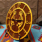 This Crooks Design round Tableau Clock features a pair of golden-toned Kokopelli figures, stylized openwork numerals, black clock hands, and a rich brown background; side view.