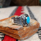 Turquoise ring by Roie Jaque featuring an oval bright blue turquoise stone in a serrated bezel, surrounded by layered, symmetrical silverwork with oxidized accents; side view.