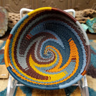Handwoven triangular wire art bowl by Bridge for Africa featuring a spiral pattern in recycled wire with slate blue, mustard yellow, charcoal, brown, and gray tones, ideal for home décor or wall display.