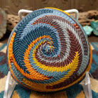 Handwoven triangular wire art bowl by Bridge for Africa featuring a spiral pattern in recycled wire with slate blue, mustard yellow, charcoal, brown, and gray tones, ideal for home décor or wall display; underside view.